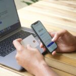 Persona realiza una transacción digital desde un teléfono junto a un computador y una tarjeta.