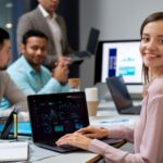 Equipo de profesionales de tecnología en una oficina; una mujer sonríe frente a un notebook que muestra un panel de datos.