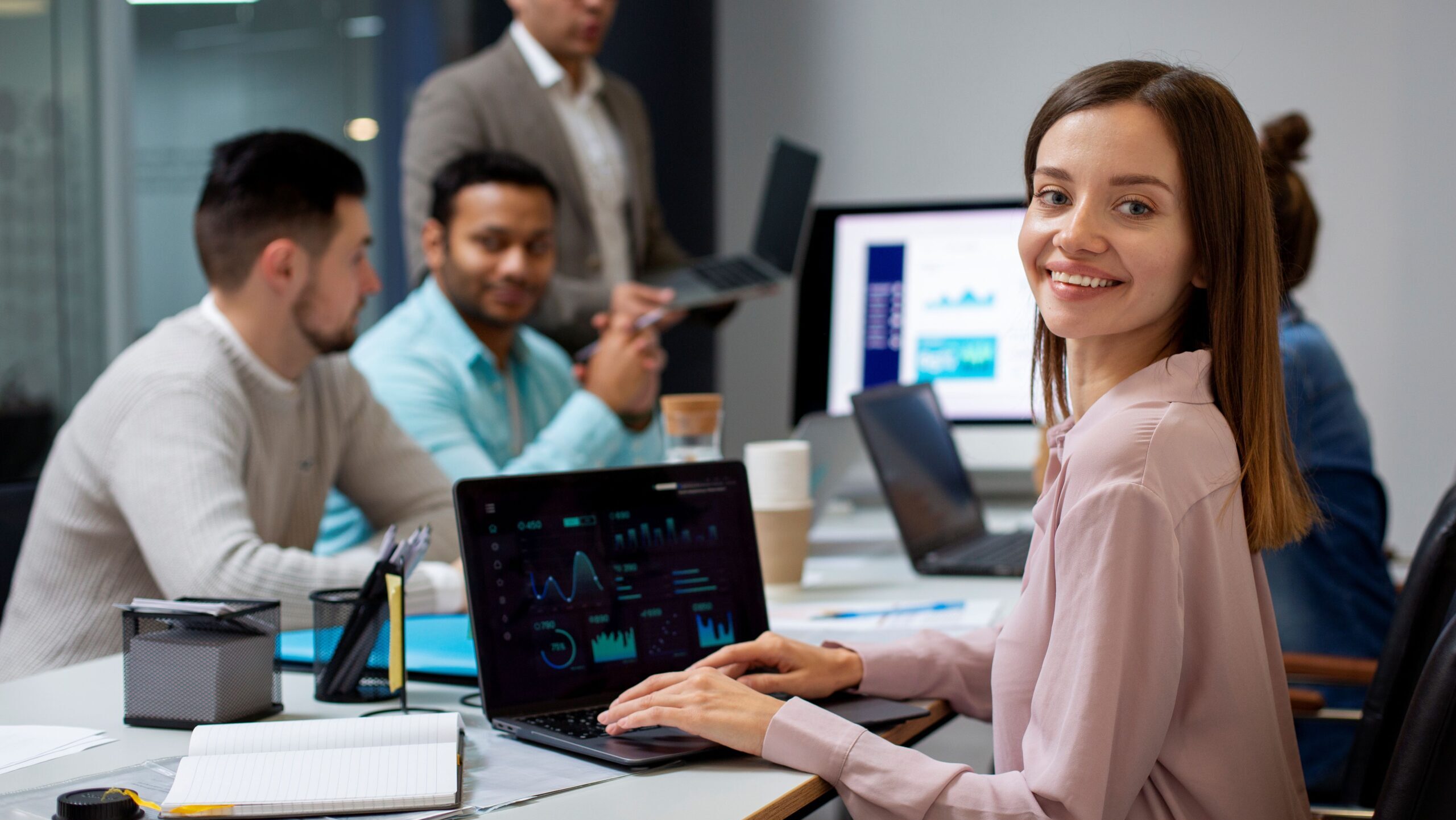 Equipo de profesionales de tecnología en una oficina; una mujer sonríe frente a un notebook que muestra un panel de datos.