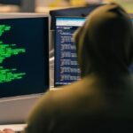 profesionales de ciberseguridad — persona con capucha frente a monitores con código