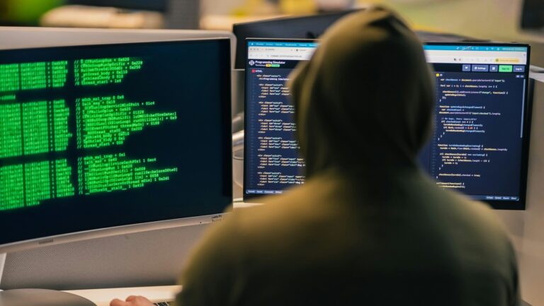 profesionales de ciberseguridad — persona con capucha frente a monitores con código